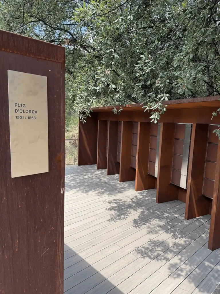 Testero de acero corten del columbario, con la galería de columbarios al fondo bajo la encina